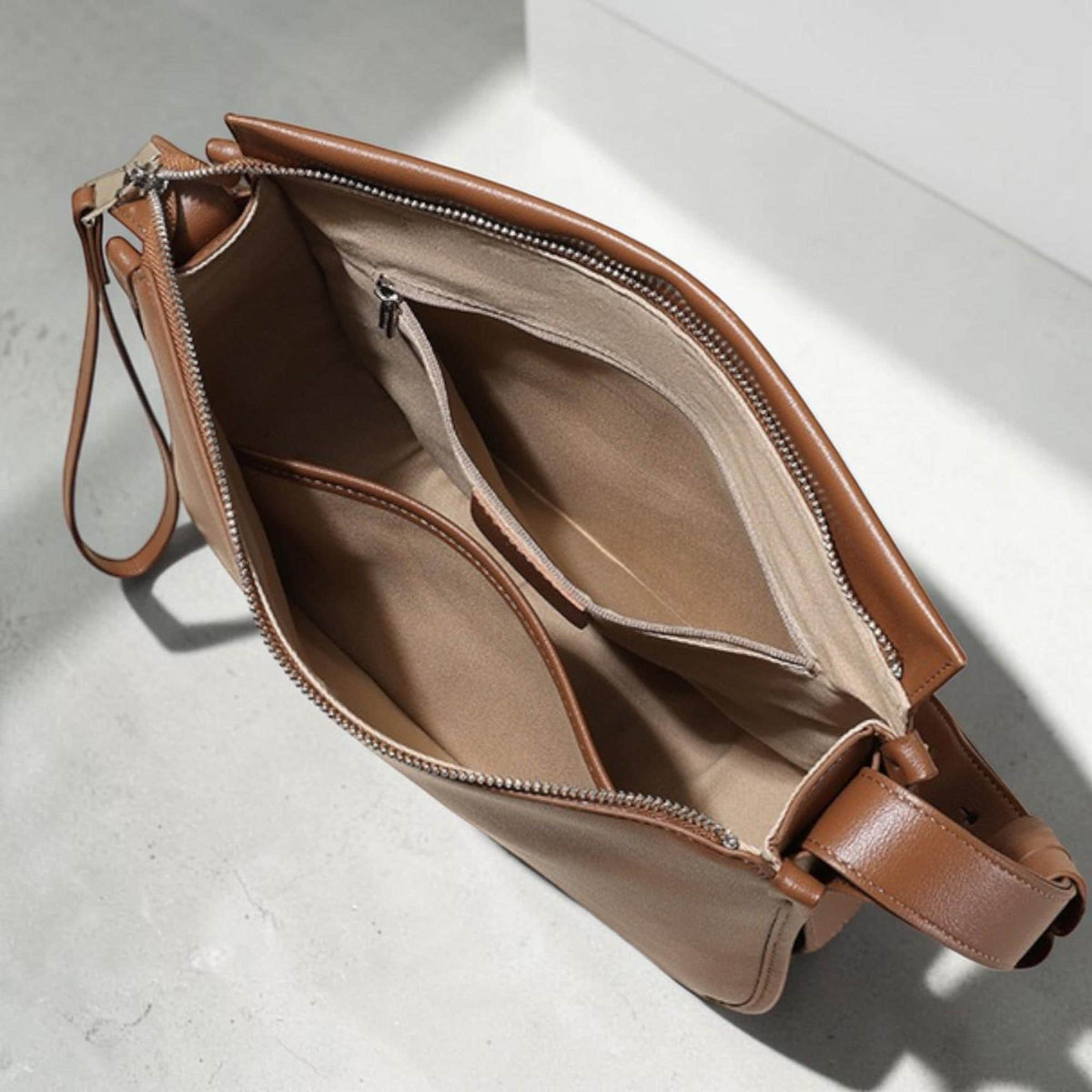 May include: Open view of a brown leather shoulder bag with multiple interior compartments. The bag has a zipper closure and a wrist strap. The interior lining is a lighter shade of brown. The bag is sitting on a light gray surface.