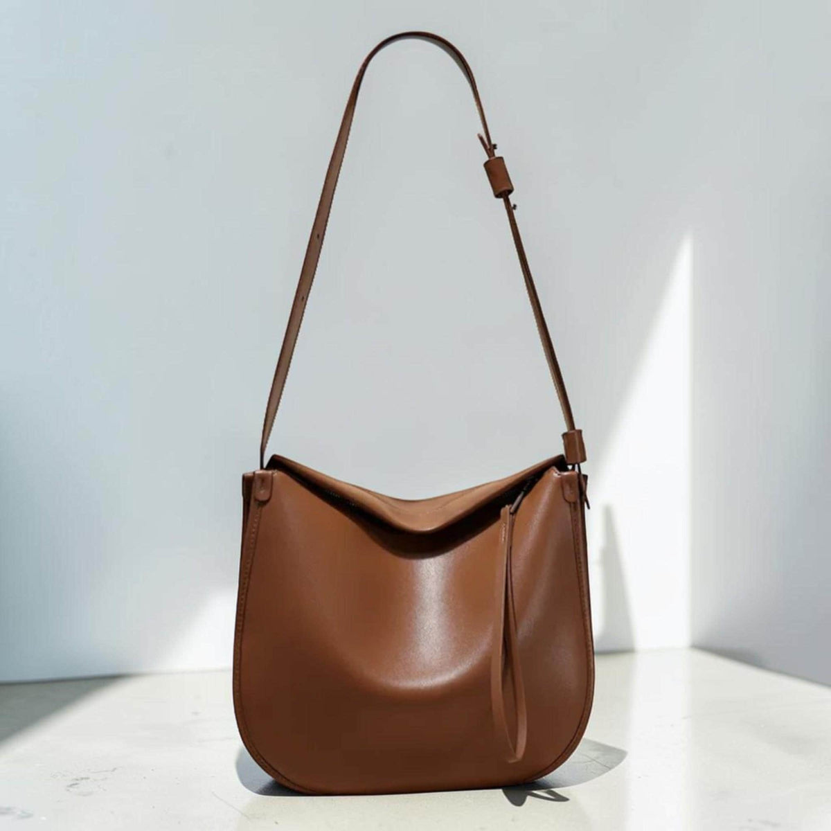 May include: A brown leather shoulder bag with a long, adjustable strap. The bag has a curved, slouchy design and a small, dangling strap detail. The bag is made of smooth leather and has a minimalist aesthetic.