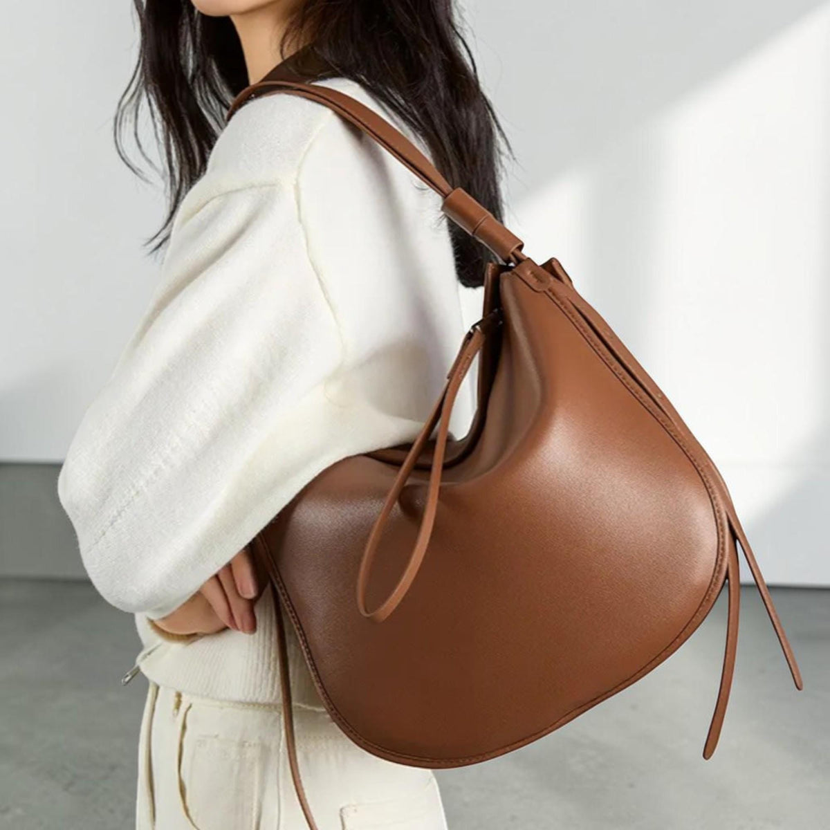 May include: A brown leather shoulder bag with a curved, crescent shape. The bag has a long strap and a drawstring closure. The bag is made of smooth leather and has a minimalist design. The bag is a medium size and appears to be empty.
