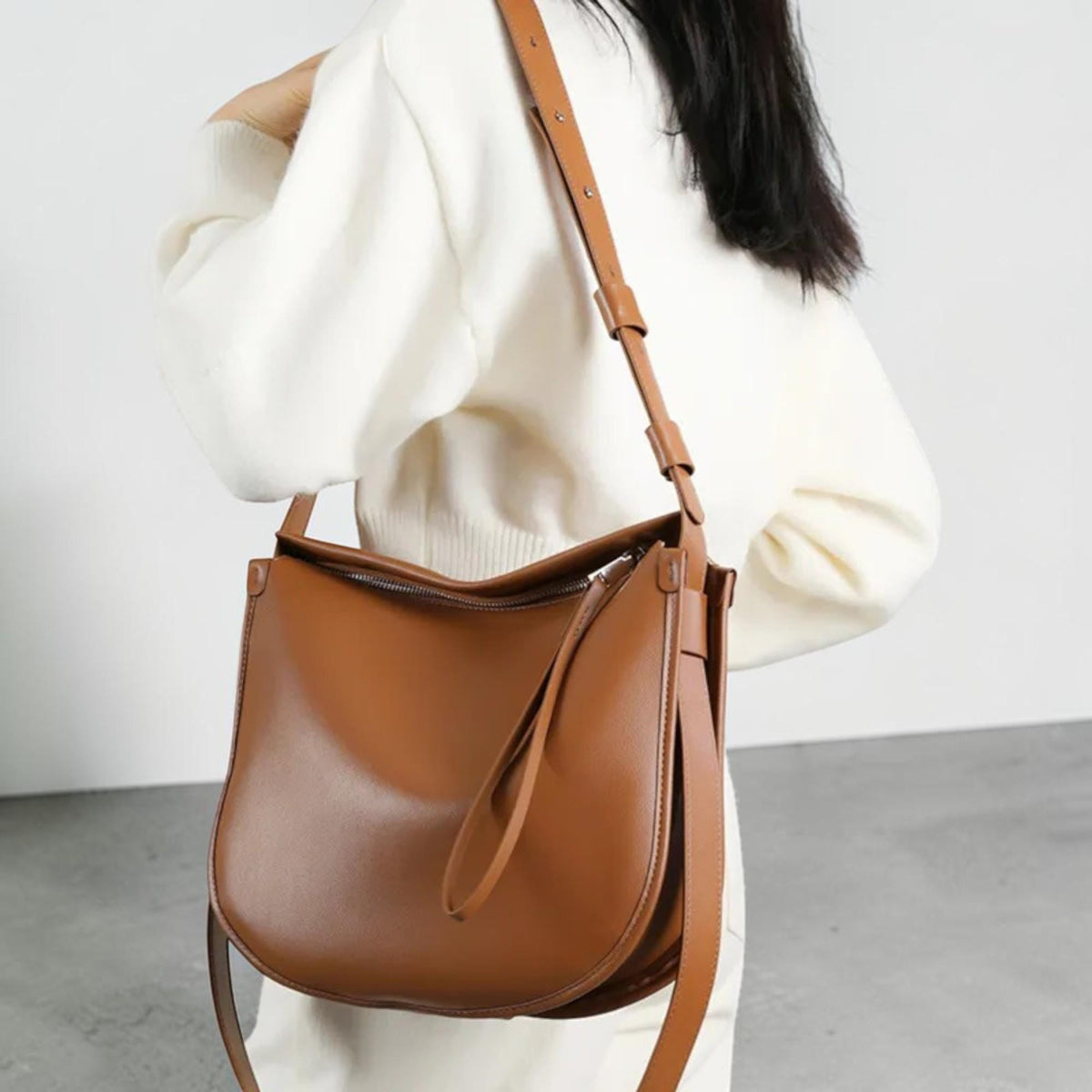 May include: A brown leather shoulder bag with a long adjustable strap. The bag has a curved bottom and a zipper closure. The strap is attached to the bag with metal hardware. The bag is worn over the shoulder.