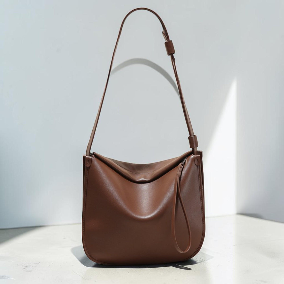May include: A brown leather shoulder bag with a long strap. The bag has a rounded shape and a simple design. The strap is adjustable. The bag is made of smooth leather and has a minimalist aesthetic.