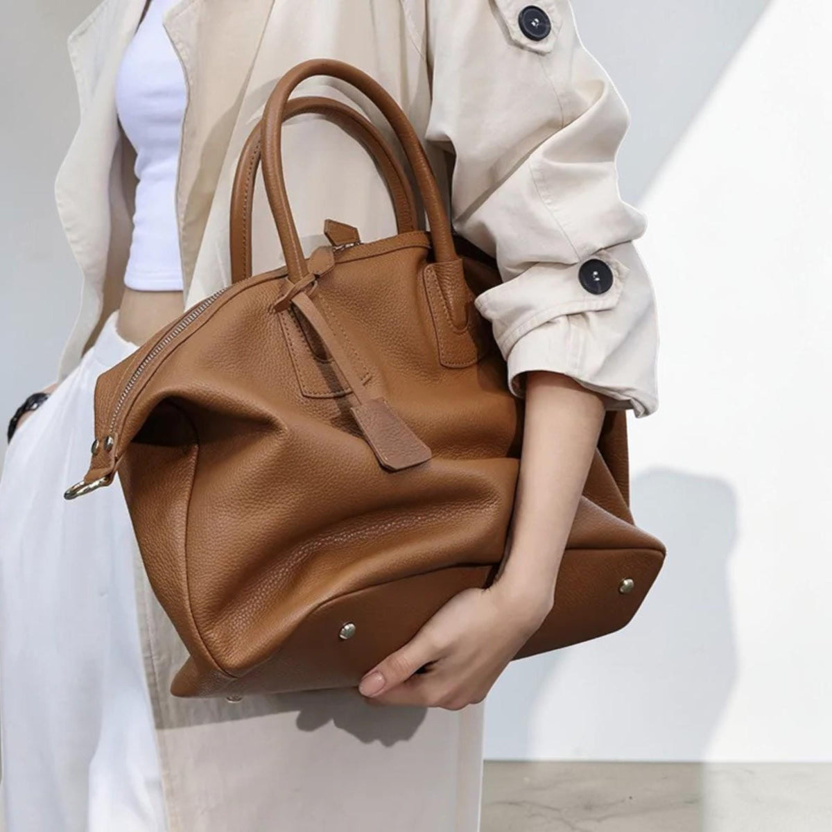 May include: A brown leather handbag with a top handle and a zipper closure. The bag has a rectangular shape with a rounded top and a small tag attached to the zipper. The bag is held by a person.