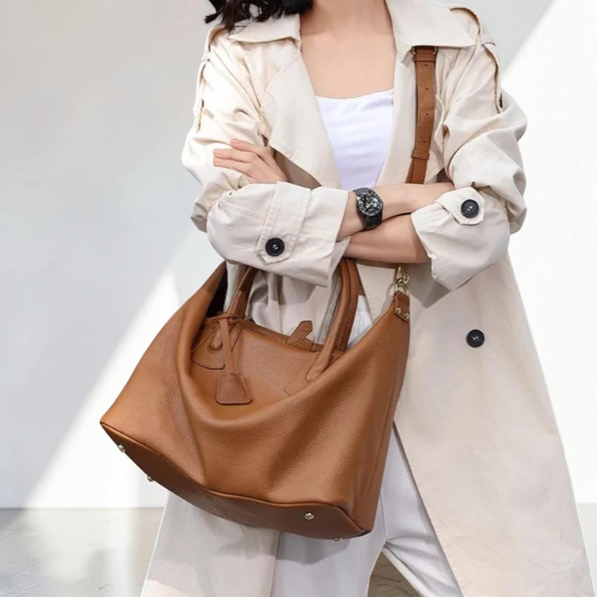 May include: A brown leather tote bag with a top handle and a shoulder strap. The bag has a soft, slouchy design and a small, decorative leather tag. The bag is carried by a person wearing a beige trench coat.