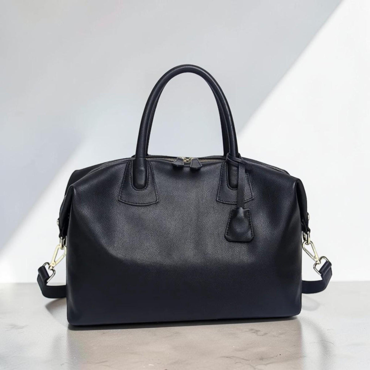 May include: A black leather duffel bag with a top handle and a detachable shoulder strap. The bag has a zipper closure and a small leather tag attached to the handle. The hardware is silver-colored.