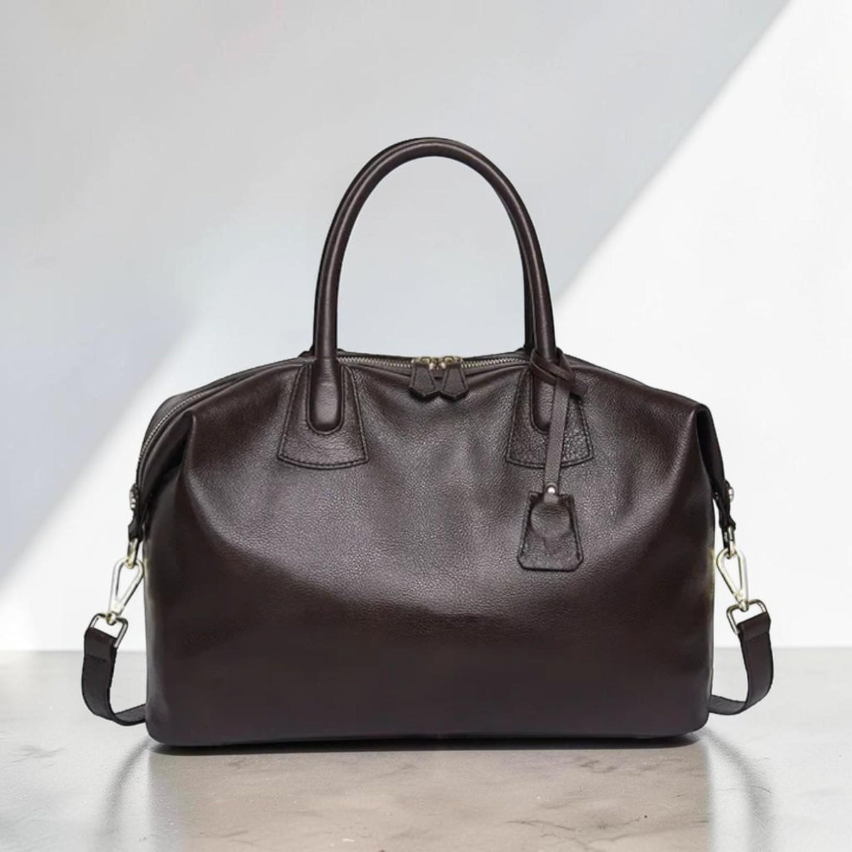 May include: A dark brown leather duffel bag with a top zipper closure, two top handles, and a detachable shoulder strap. The bag has a small leather tag attached to the zipper. The hardware is silver-colored.