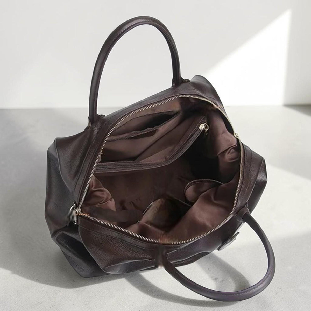 May include: Open brown leather handbag with two handles and a zipper closure. The interior is visible, revealing pockets and a brown lining. The bag is sitting on a light gray surface.