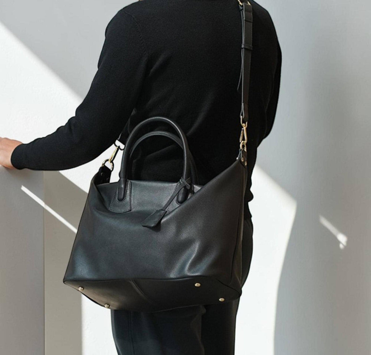 May include: A black leather tote bag with a top handle and a detachable shoulder strap. The bag has a rectangular shape with a trapezoidal side profile. Gold-colored hardware accents the bag's design.