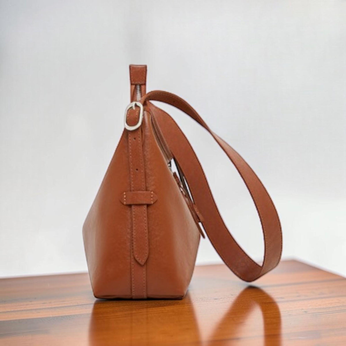May include: A brown leather shoulder bag with a silver-tone zipper and buckle. The bag has a long, adjustable strap and a structured, geometric shape. The leather appears to be smooth and the bag is empty.