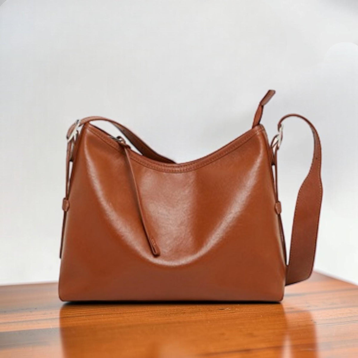 May include: A brown leather shoulder bag with a curved top and adjustable strap. The bag has a smooth, glossy finish and a long, thin strap that can be worn over the shoulder or across the body. The bag is sitting on a wooden surface.