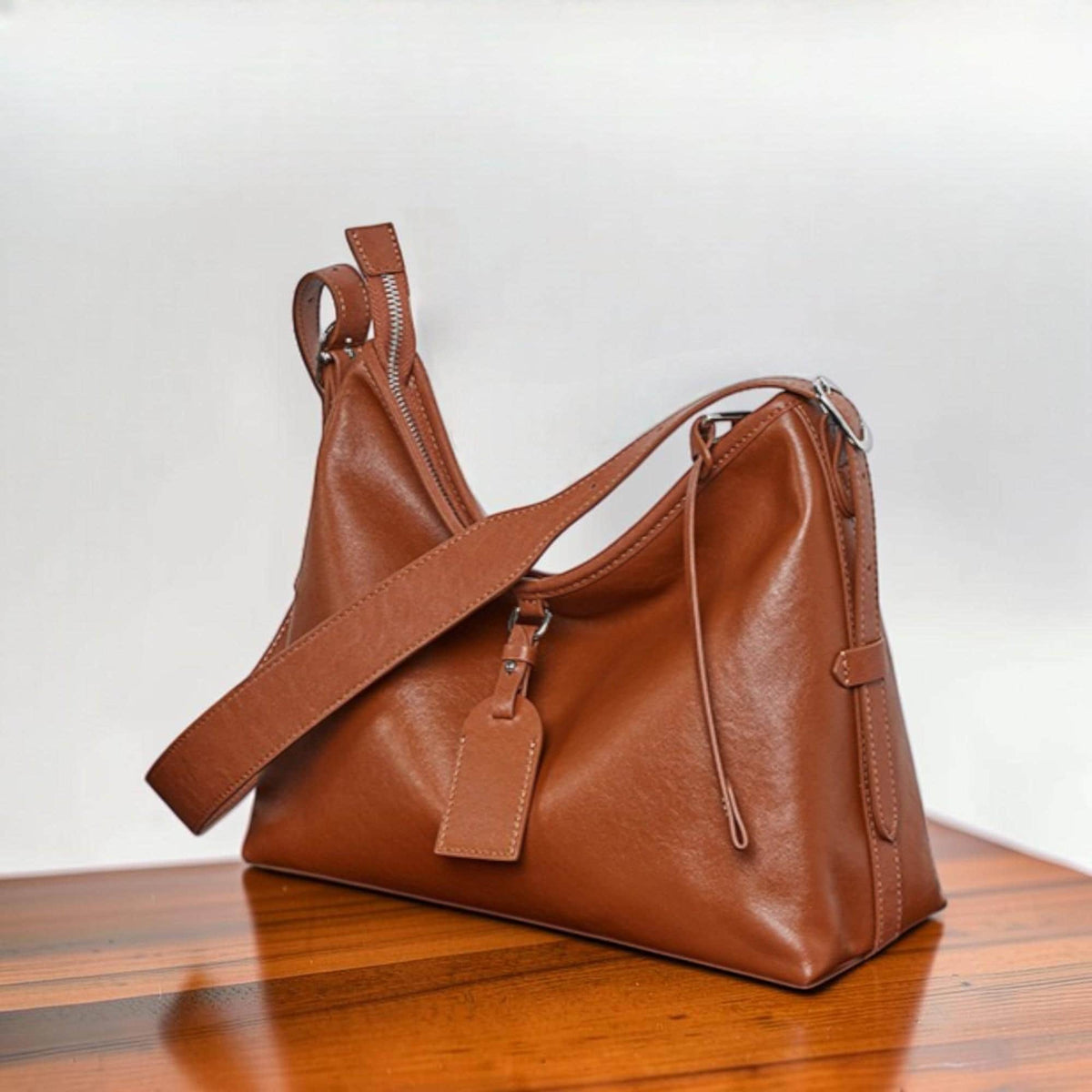 May include: A brown leather shoulder bag with a long adjustable strap and a smaller handle. The bag has a rectangular shape with a zipper closure and a small tag attached to the strap. The bag is sitting on a wooden surface.