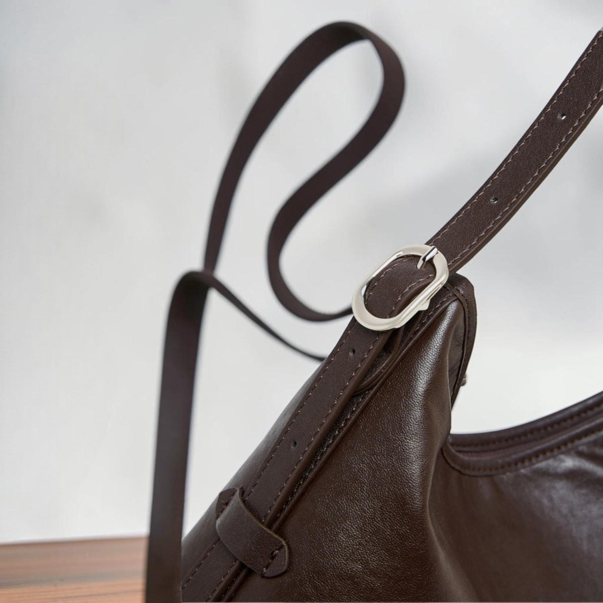 May include: Close-up of a brown leather shoulder bag with a long strap. The bag features a silver-colored buckle and a smaller strap detail. The leather appears smooth and the bag has a curved shape.