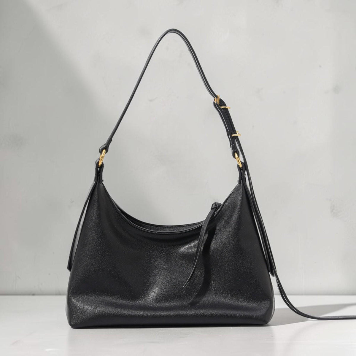 May include: A black shoulder bag with a curved shape and a long, adjustable strap. The bag has a smooth texture and gold-colored hardware accents. The strap is attached to the bag with gold-colored rings.