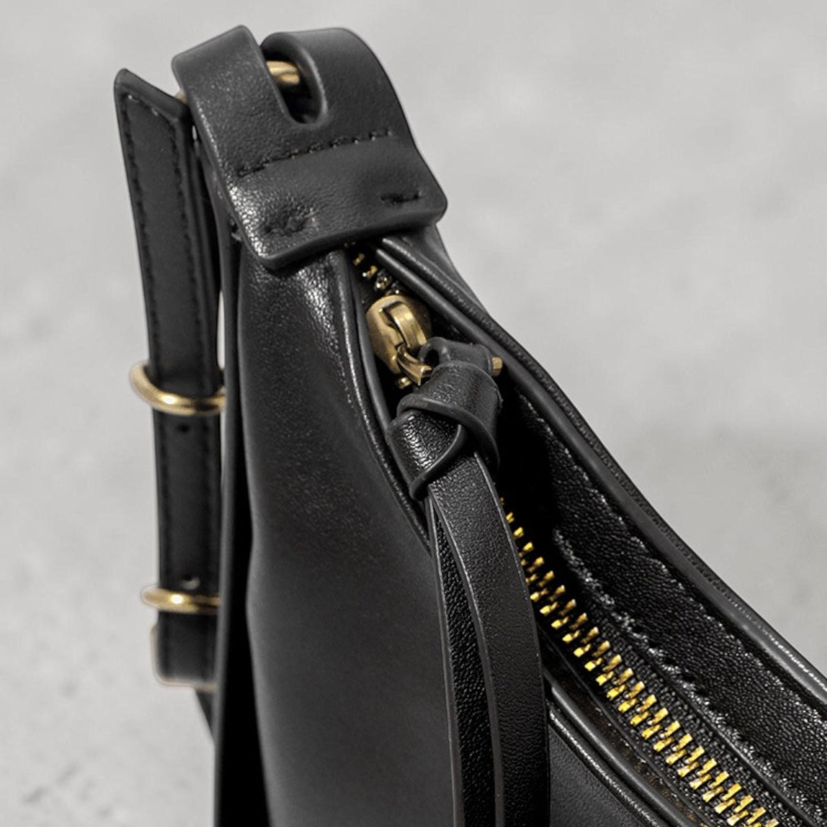 May include: Close-up of a black leather shoulder bag. The bag features a gold-colored zipper and hardware, with a strap and a small leather pull. The bag's design is minimalist and sleek, suitable for everyday use.
