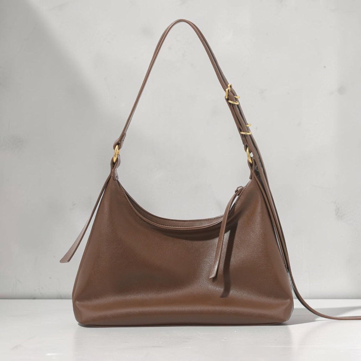 May include: A brown leather shoulder bag with a curved design. The bag has a long strap and gold-colored hardware. The bag is made of a smooth, solid material and has a simple, elegant style.