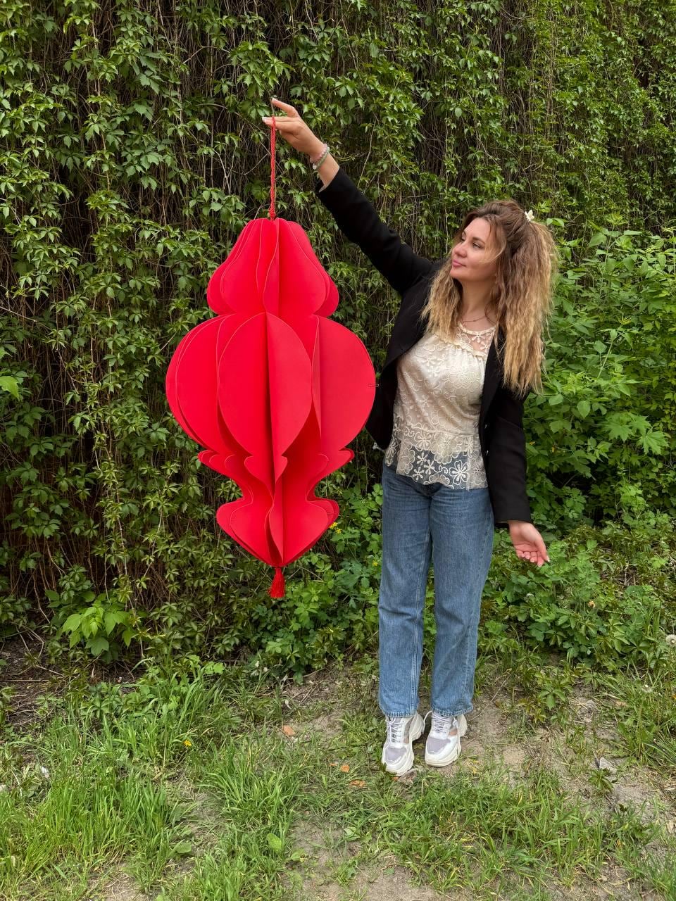 May include: A large, vibrant red paper lantern with a layered design is suspended from a red string. The lantern has a bulbous shape with decorative cutouts and a tassel at the bottom. The background is a lush green wall of foliage.