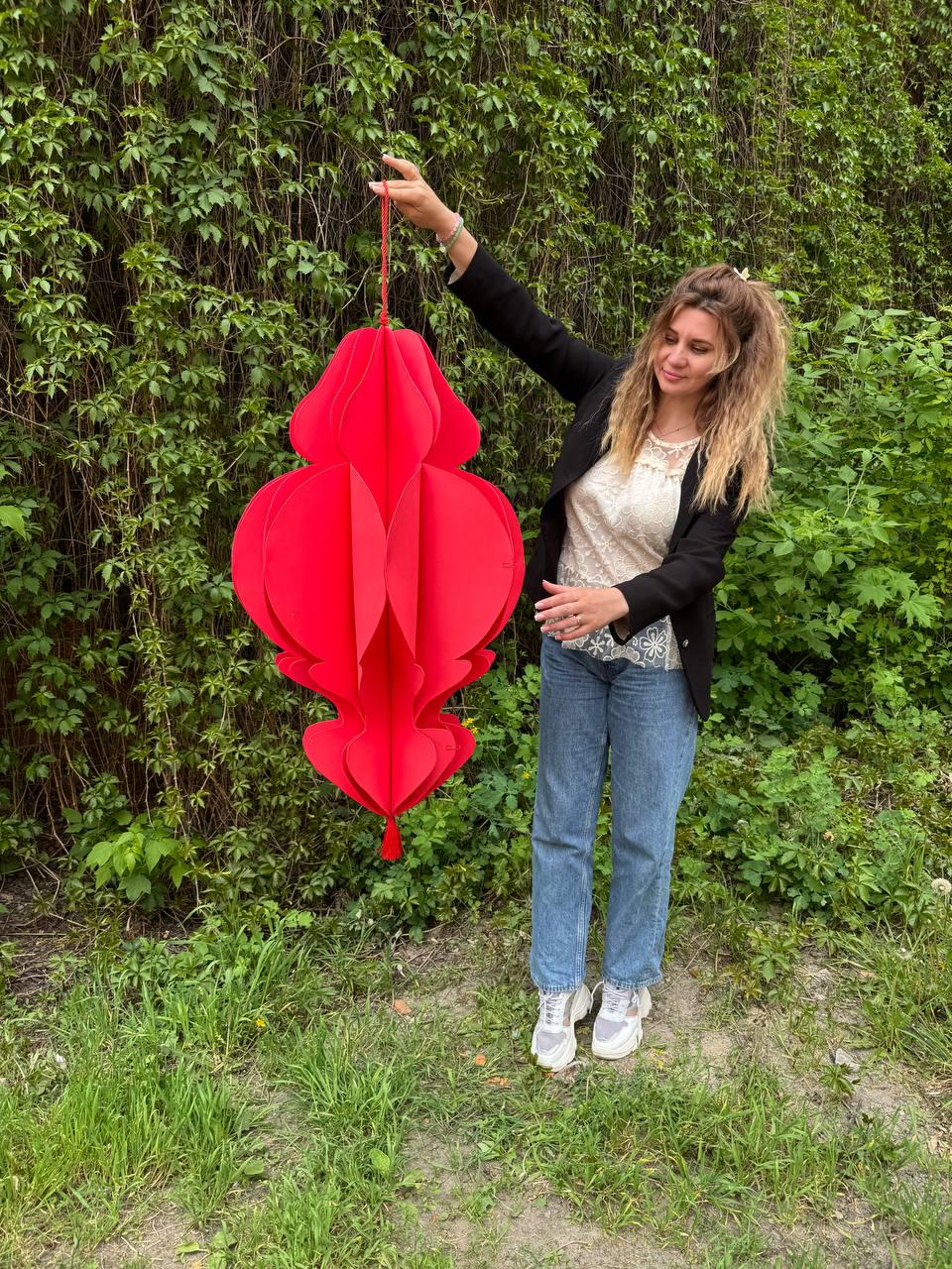 May include: A large, vibrant red paper lantern with a layered, floral-like design. The lantern is suspended by a red string and features a tassel at the bottom. The lantern is held up against a backdrop of green foliage.