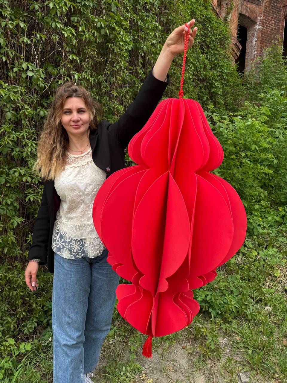 May include: A large, vibrant red paper lantern with a layered, petal-like design. The lantern is suspended by a red cord and tassel. The overall shape is bulbous, with a decorative, scalloped bottom. The background features lush green foliage.