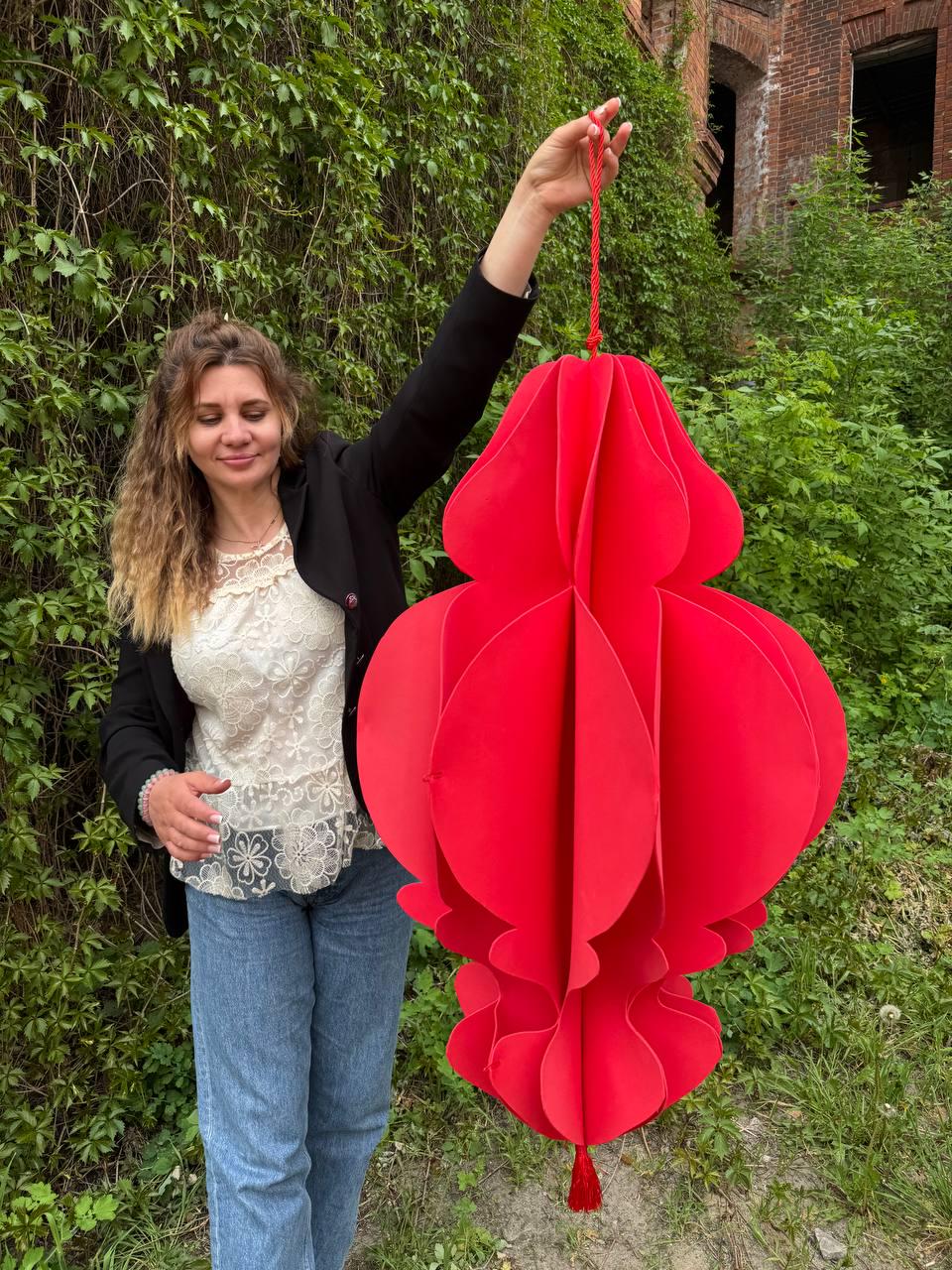 May include: A large, vibrant red paper lantern with a layered, floral-like design. The lantern is suspended by a red cord and tassel. The decorative piece is held up against a backdrop of green foliage and a brick building.
