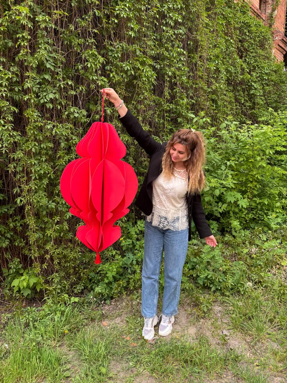 May include: A large, vibrant red paper lantern with a layered, ornate design. The lantern is suspended by a red string and held up against a backdrop of lush green foliage. The lantern's shape is reminiscent of a traditional Chinese lantern.