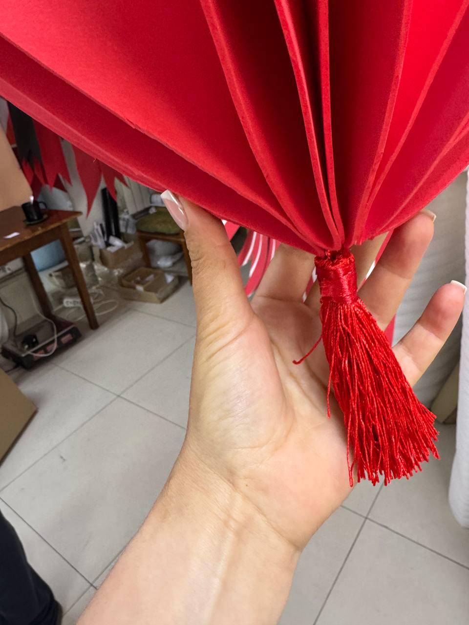 May include: A bright red paper lantern with a tassel, held in a hand. The lantern is made of layered, fan-like sections. The tassel is made of red threads. The background is a light-colored tiled floor.