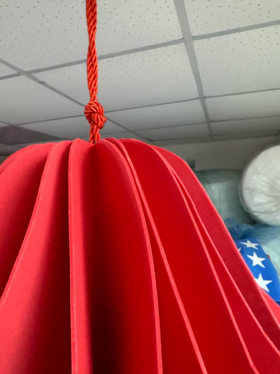 May include: A vibrant red paper lantern, suspended from a matching rope. The lantern has a layered, pleated design, creating a unique, eye-catching shape. The rope is knotted at the top, securing the lantern for hanging.