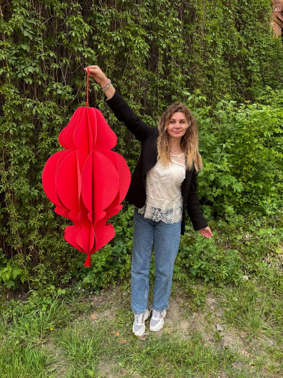 May include: A large, vibrant red paper lantern with a layered, ornate design. The lantern is suspended by a red string. The lantern is held up in front of a green leafy background.