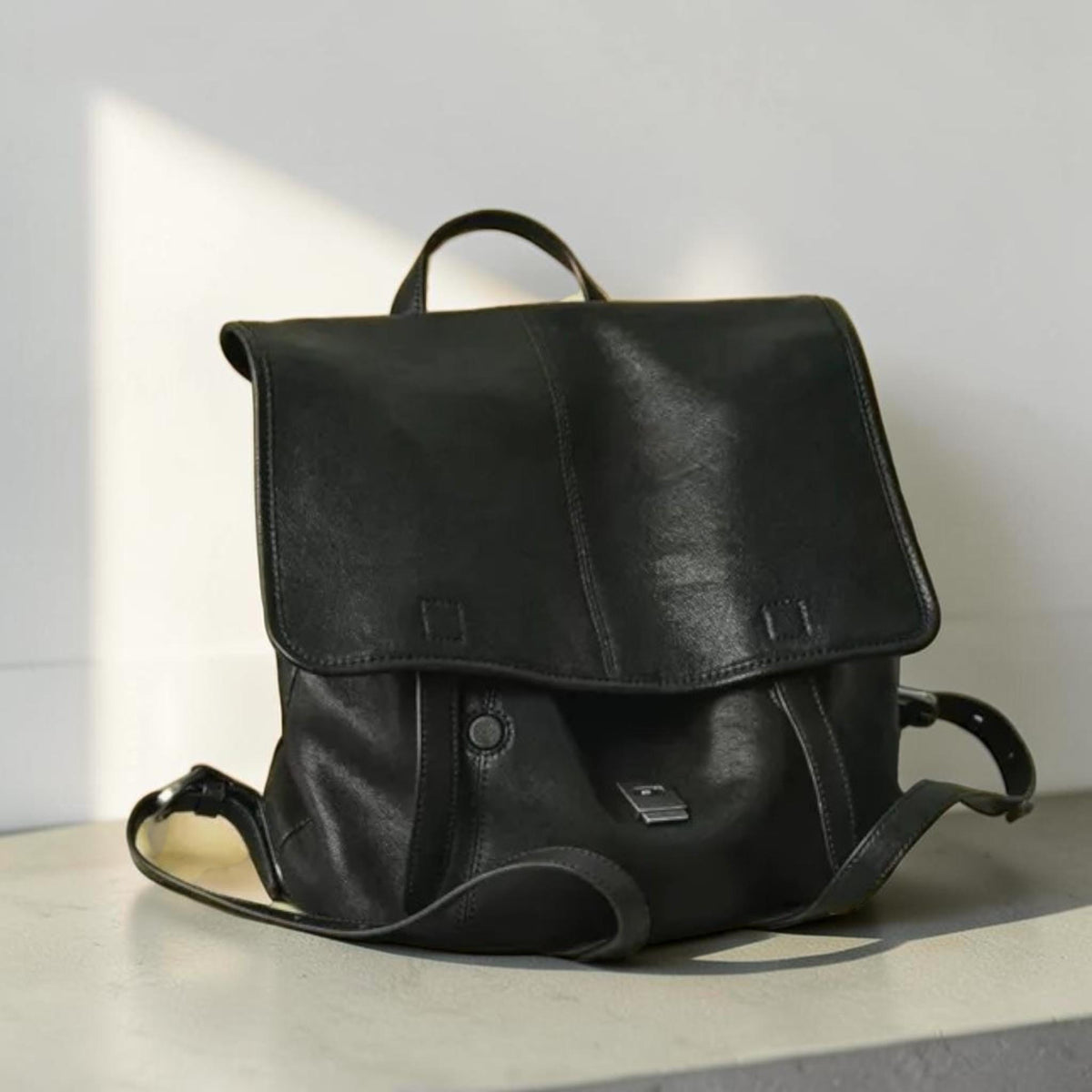 May include: A black leather backpack with a flap closure and adjustable straps. The bag has a top handle and a small metal accent on the front. The leather appears to be slightly textured, and the bag is sitting on a light-colored surface.