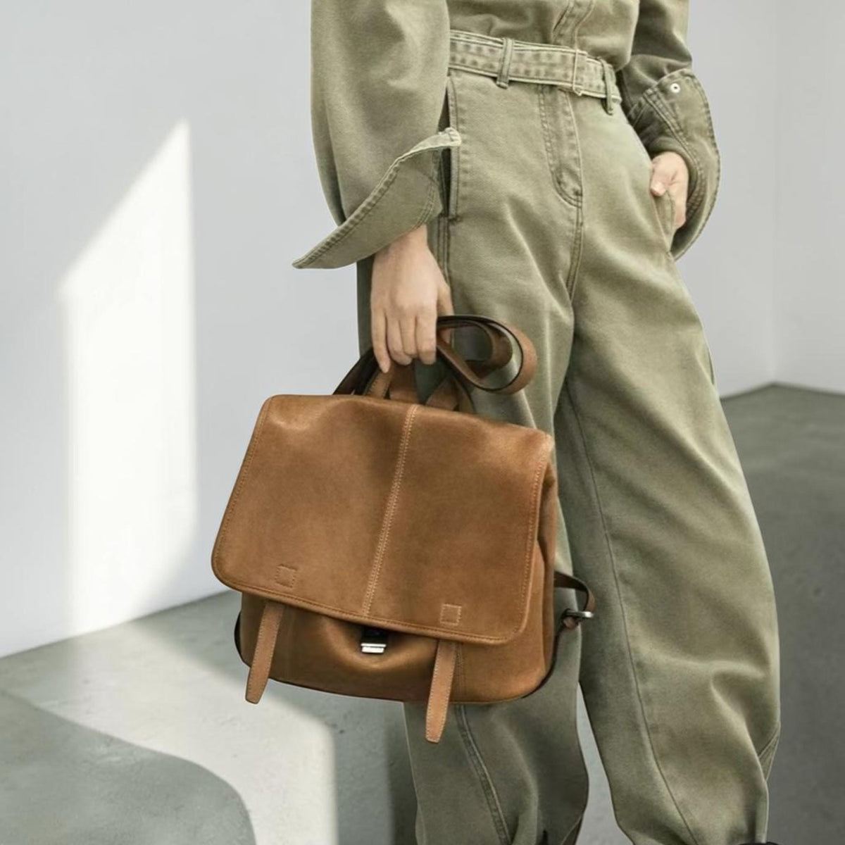 May include: A brown leather backpack with a flap closure, two front straps, and a top handle. The bag has a rectangular shape with a vertical seam down the center. The backpack is held by a person wearing olive green pants and a matching jacket.