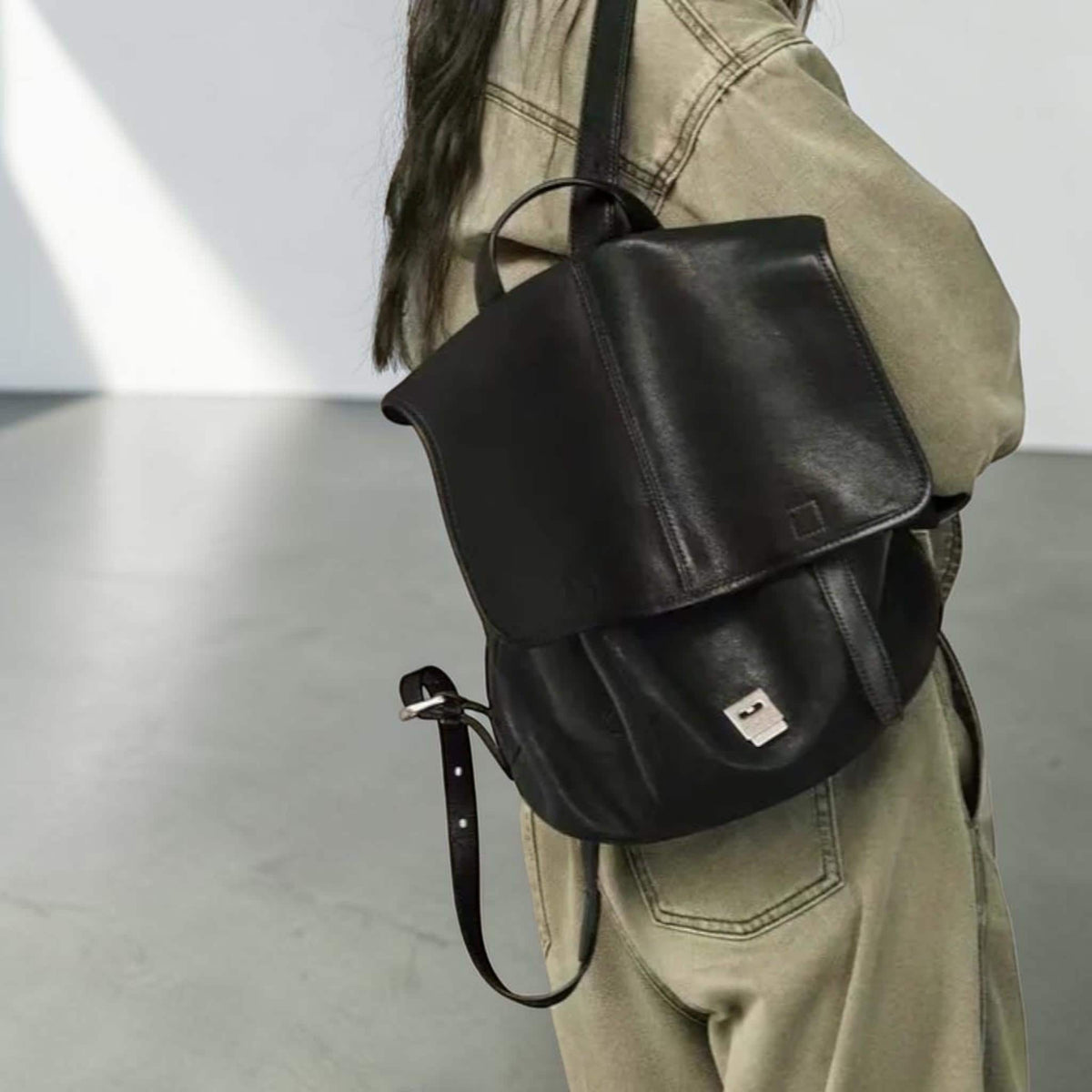 May include: Black leather backpack with a flap closure and a top handle. The backpack has a square shape with a silver buckle on the strap. The bag is worn over the shoulder.