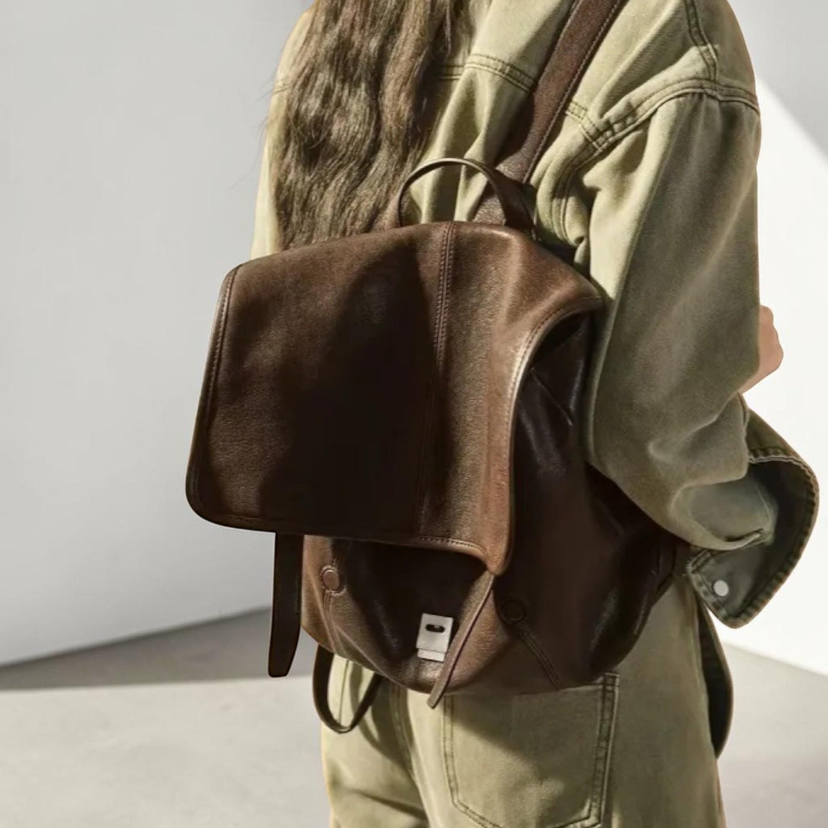 May include: A brown leather backpack with a flap closure and a top handle. The backpack has adjustable straps and a silver-colored buckle. The bag is worn on the back.