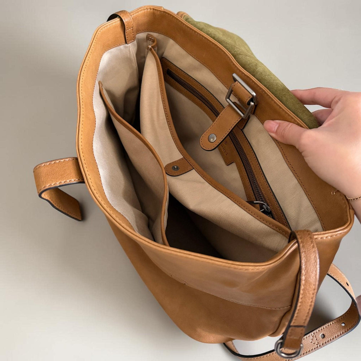 May include: Open view of a tan leather shoulder bag with multiple interior compartments. The bag features a brown leather strap, a silver buckle, and a green suede accent. The interior is lined with a light beige fabric.