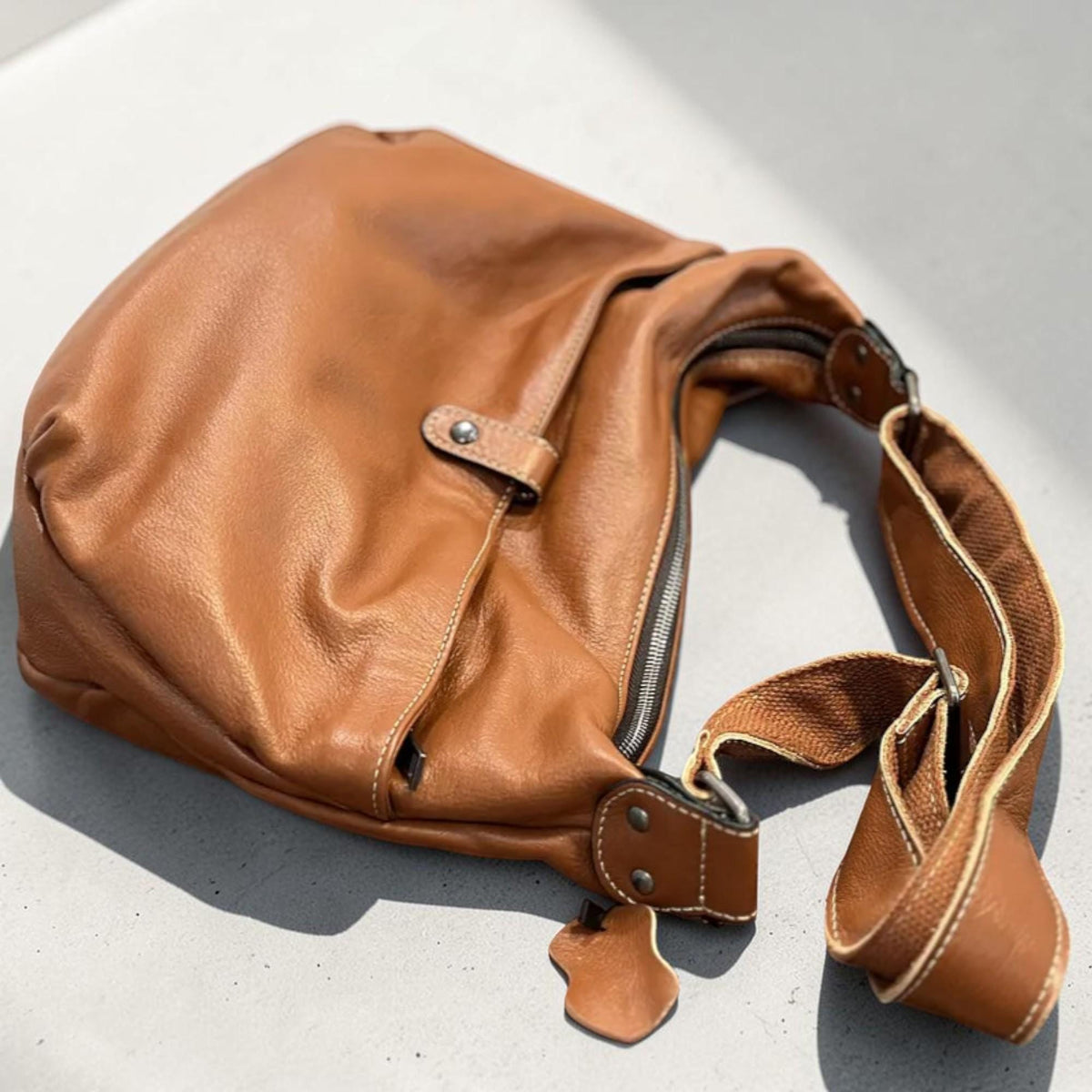 May include: A brown leather crossbody bag with a curved shape. The bag features a zipper closure, a long adjustable strap, and a small exterior pocket. The leather has a natural, textured appearance.
