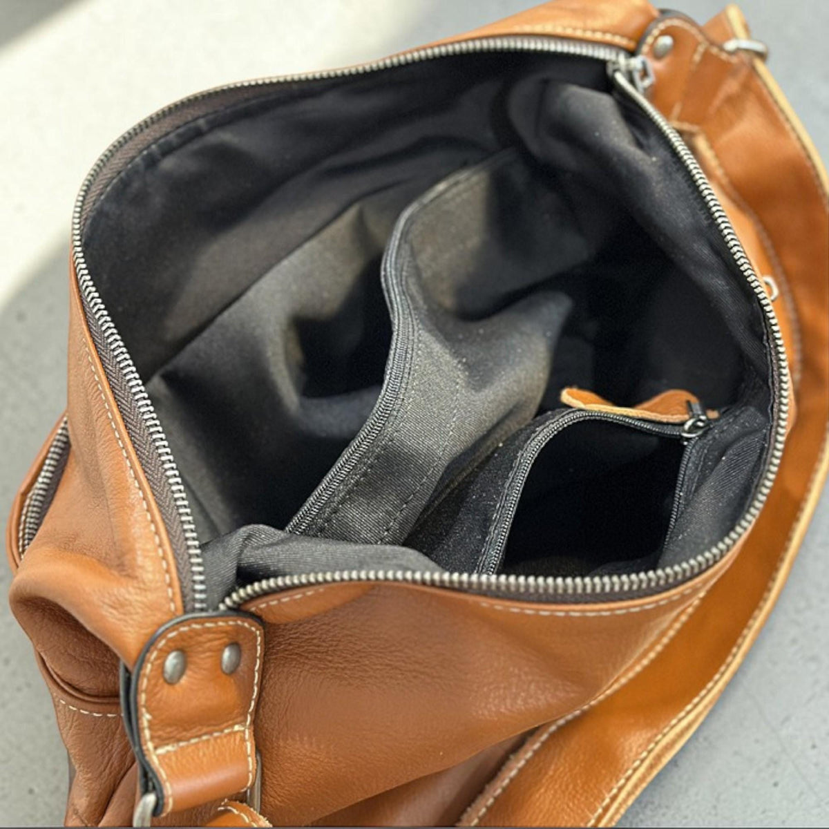 May include: Open view of a brown leather shoulder bag with a black interior. The bag has multiple compartments, including a zippered pocket. The zipper is silver-colored, and the bag has visible stitching.