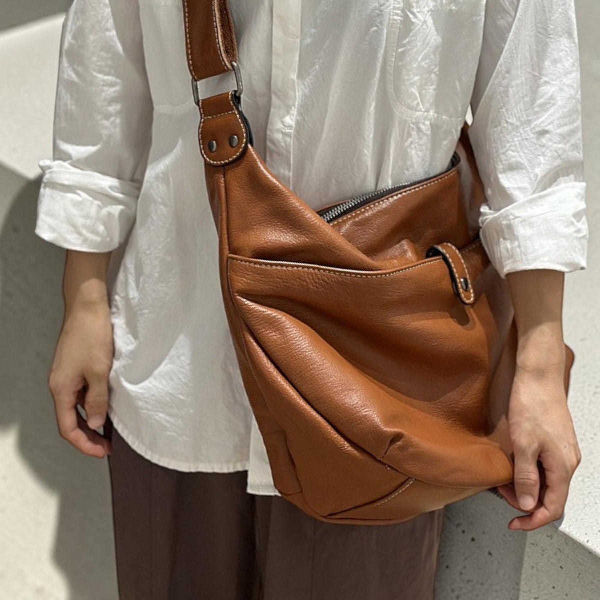 May include: A brown leather shoulder bag with a large front pocket and a zipper closure. The bag has a wide strap and is worn across the body. The bag is made of a soft, supple leather and has white stitching.