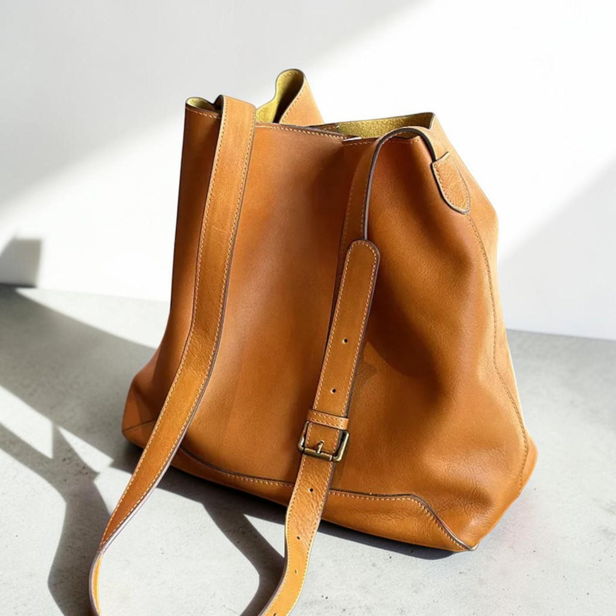 May include: A tan leather tote bag with a long shoulder strap and a smaller adjustable strap with a metal buckle. The bag has a simple, classic design with visible stitching.