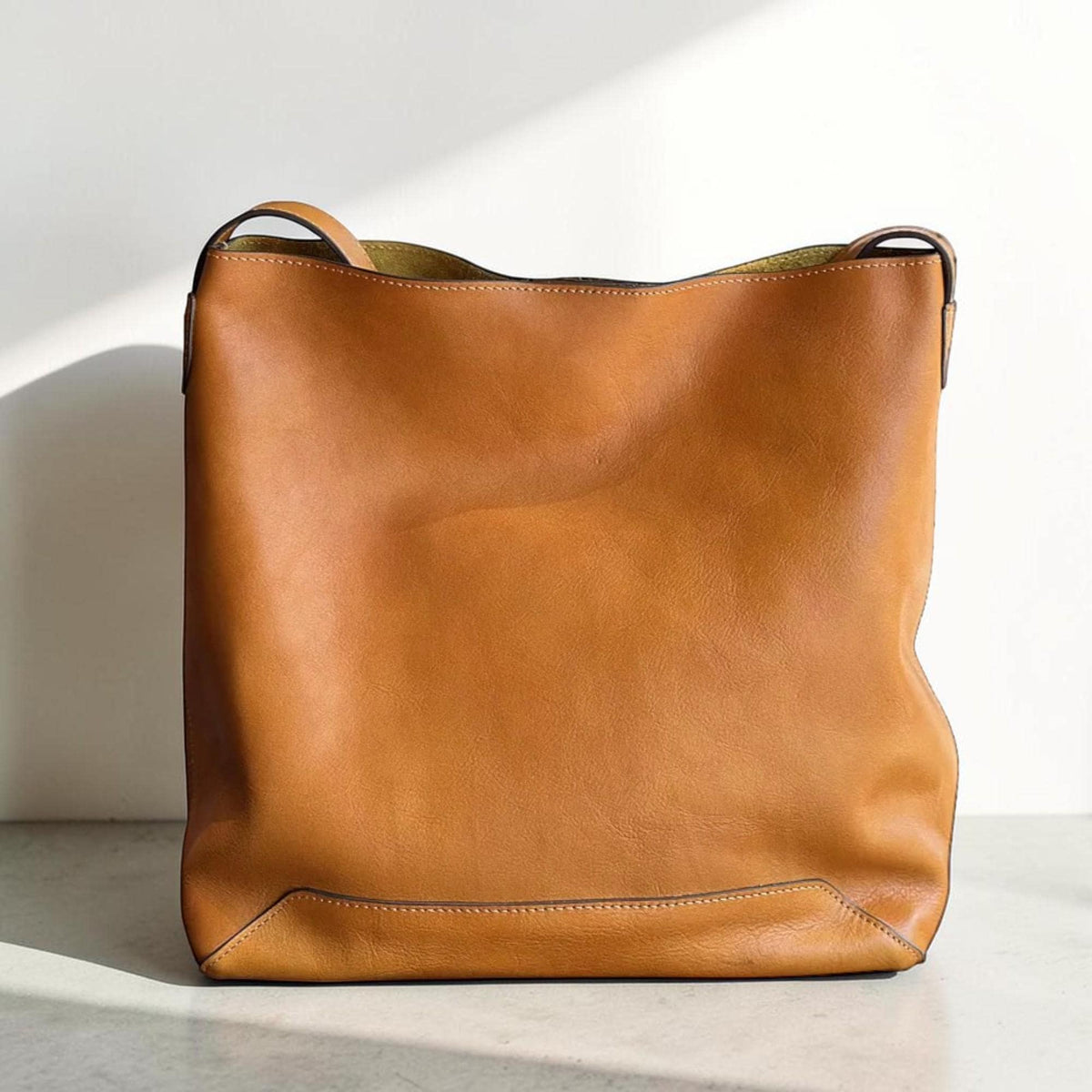 May include: A large, tan leather tote bag with a rectangular shape and a single handle. The bag has a smooth texture and a subtle sheen, with visible stitching along the bottom and sides. The bag is empty and stands upright.