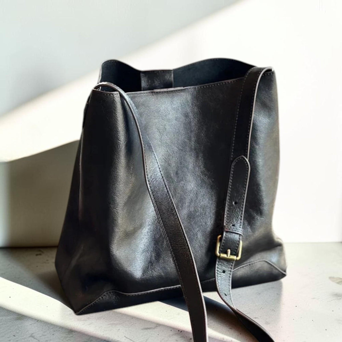 May include: A black leather bucket bag with a long adjustable strap and a gold-colored buckle. The bag has a simple, minimalist design and a spacious interior. The bag is made of a smooth, high-quality leather.