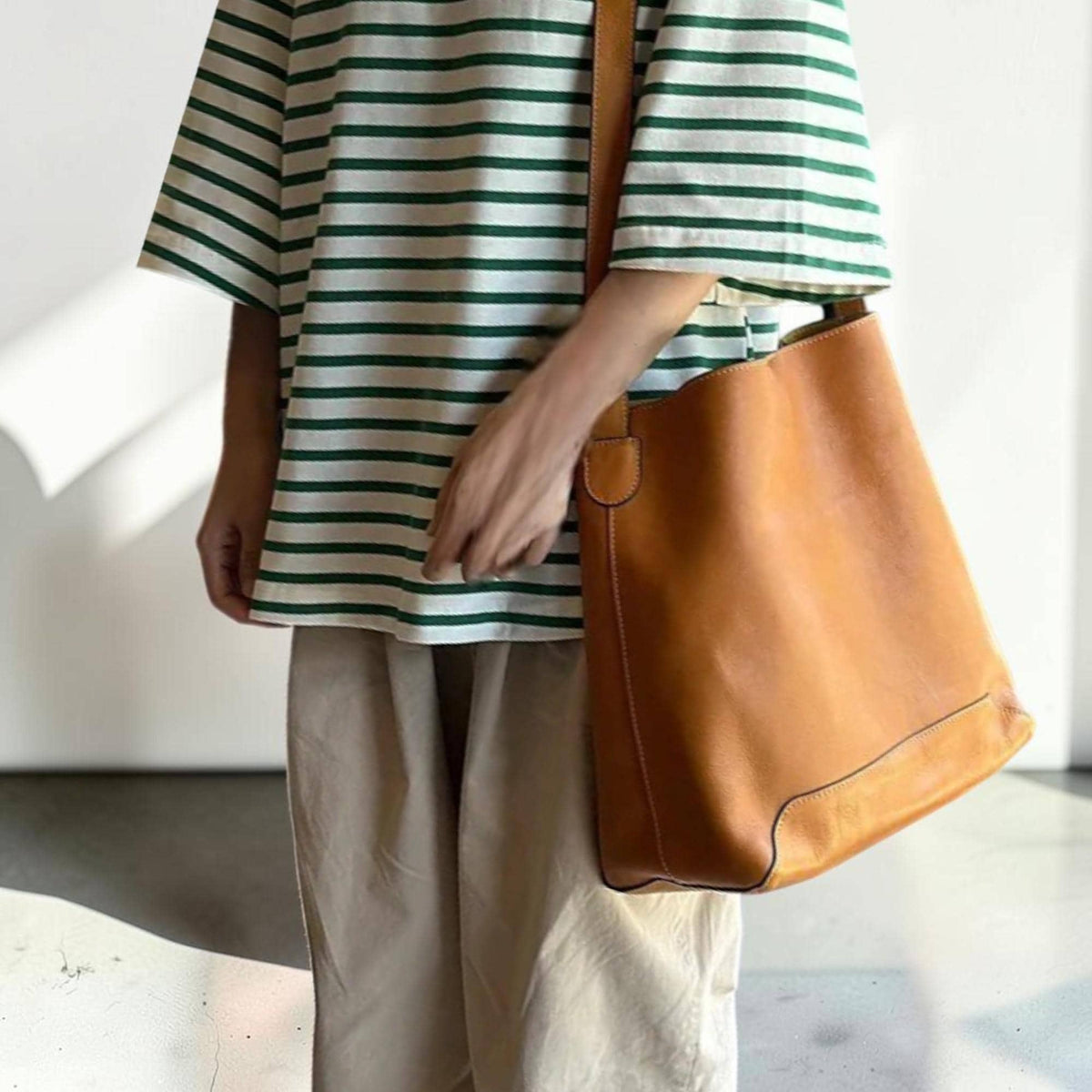 May include: A tan leather tote bag with a long shoulder strap. The bag is a light brown color and has a rectangular shape. The person is wearing a green and white striped t-shirt and tan pants.