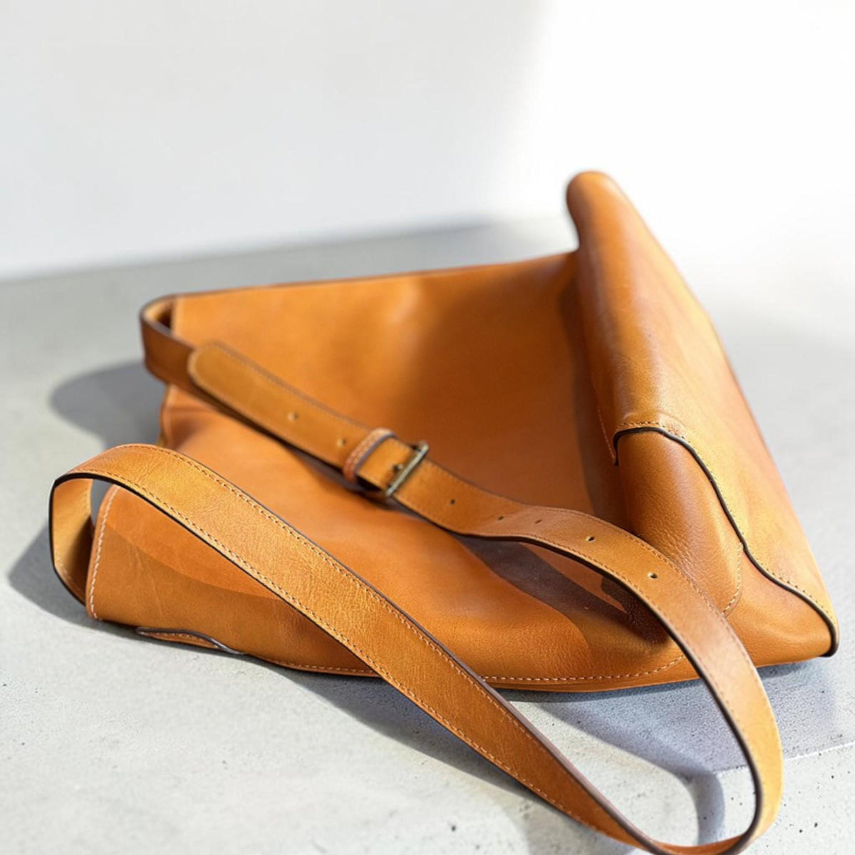 May include: A tan leather shoulder bag with a long adjustable strap and a metal buckle. The bag is made of smooth leather and has a simple, minimalist design. The bag is shown on a light gray surface.