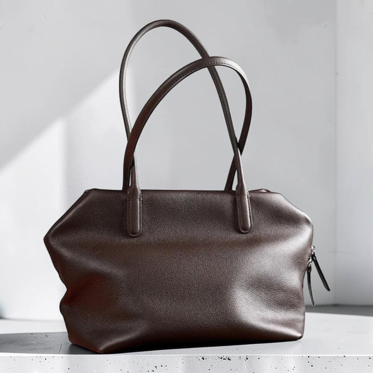 May include: A brown leather tote bag with two curved handles. The bag has a rectangular shape with a zipper closure and a textured surface. The handles are long and arched, suitable for carrying over the shoulder.