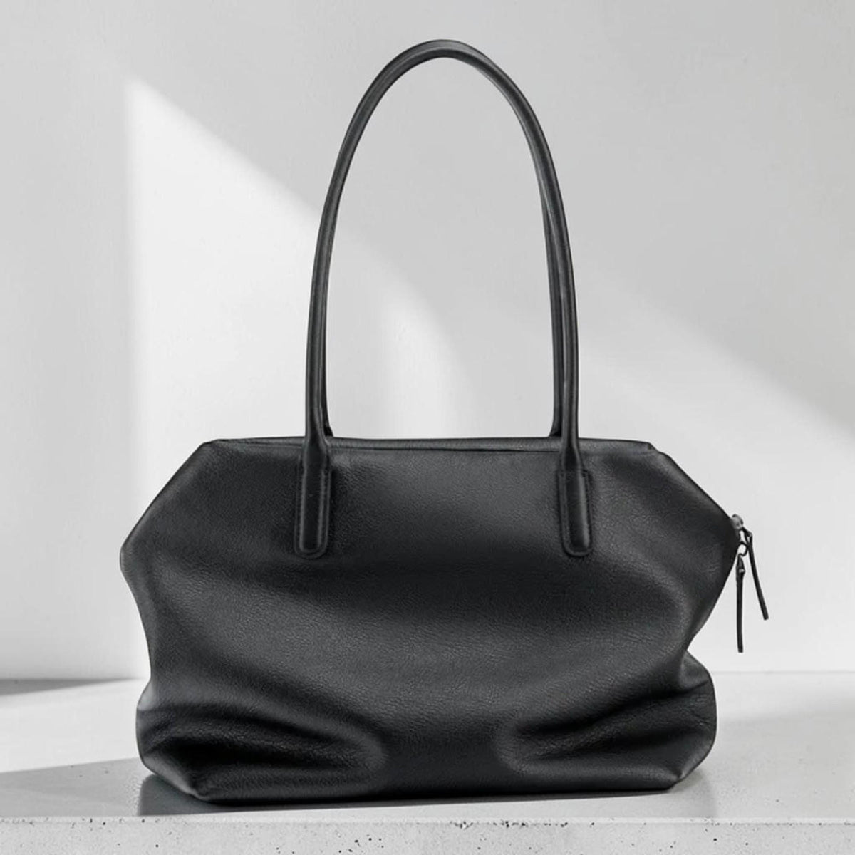 May include: A black leather tote bag with two long handles. The bag has a rectangular shape with a zipper closure and a textured surface. The bag is sitting on a white surface.