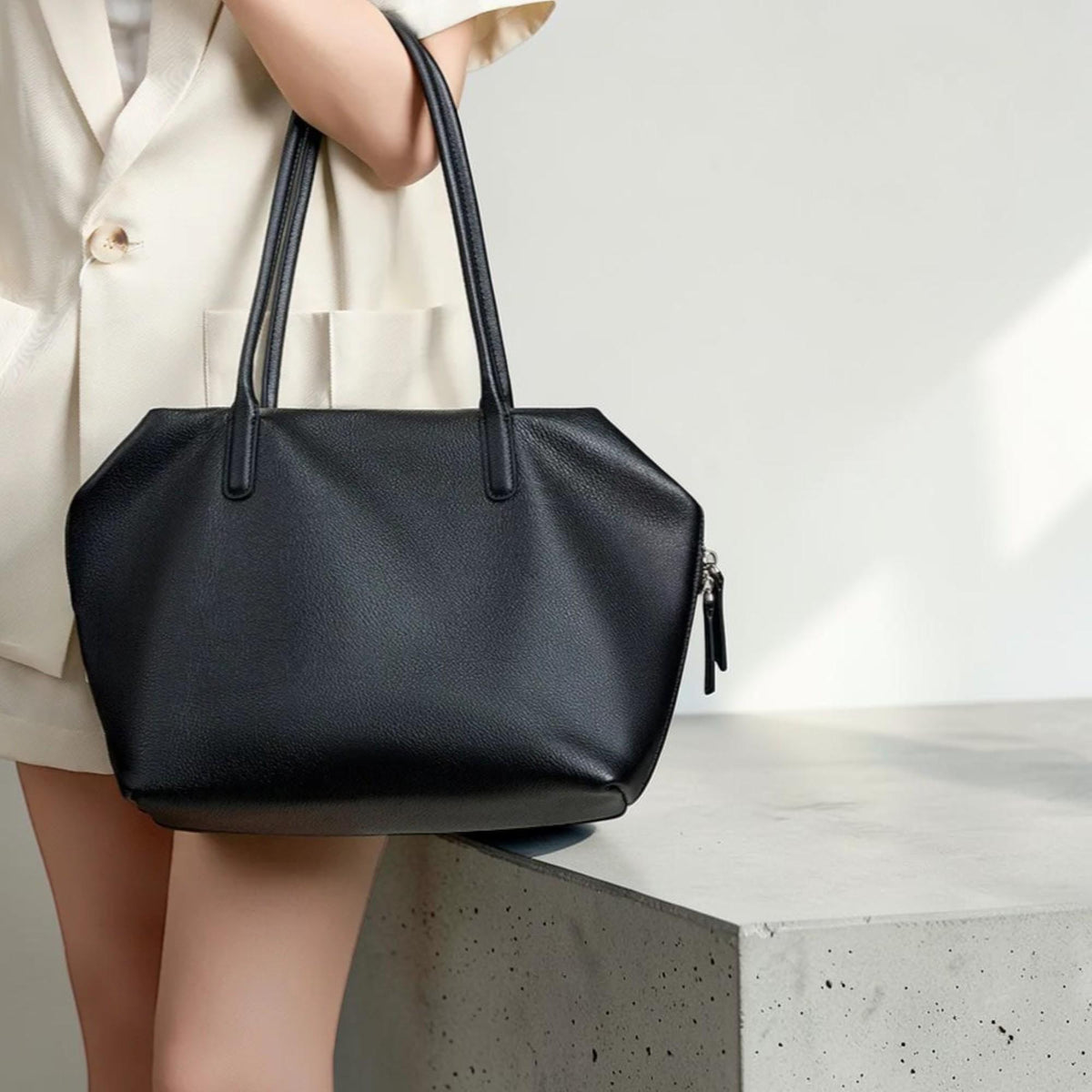 May include: A black tote bag with long handles. The bag has a geometric shape with a zipper closure on the side. The bag is held by a person wearing a cream-colored blazer. The bag is made of a smooth material.