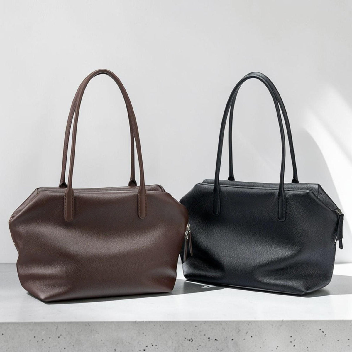 May include: Two leather tote bags, one in dark brown and the other in black, are displayed side-by-side. Each bag has a rectangular body with a zipper closure and long, slender handles. The bags are placed on a light-colored surface against a white backdrop.