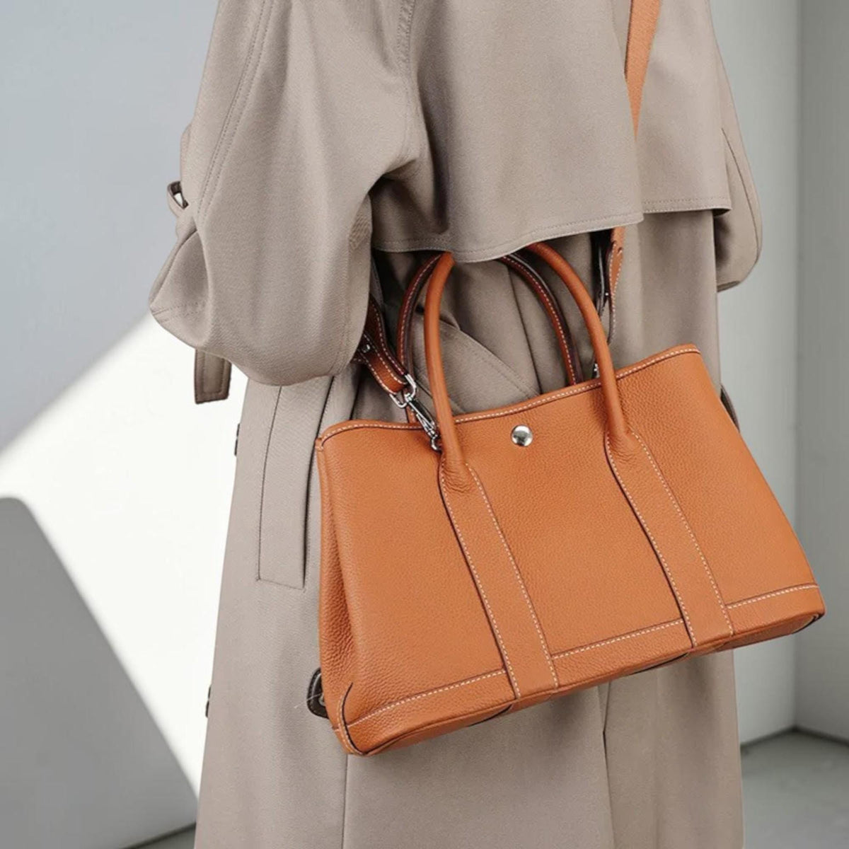 May include: A tan leather tote bag with a top handle and a shoulder strap. The bag is a warm orange-brown color with white stitching and a silver-colored clasp. The bag is being carried by someone wearing a tan trench coat.