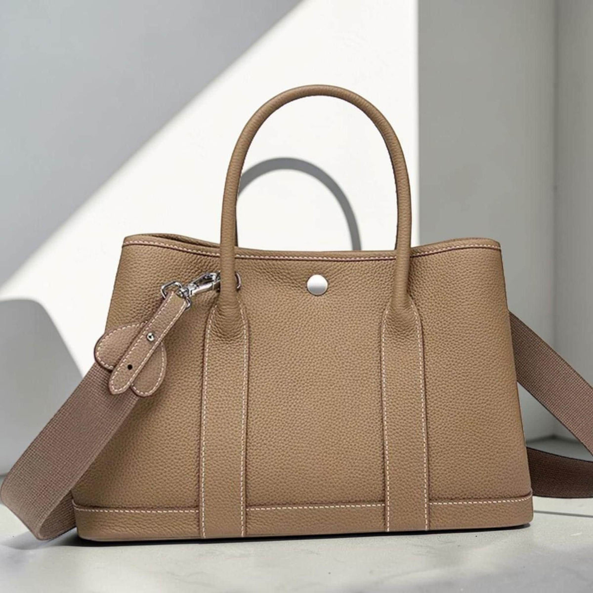 May include: A tan leather tote bag with a top handle and a detachable shoulder strap. The bag features a silver-tone clasp, a decorative leather accent, and white stitching details. The bag is designed with a structured silhouette.