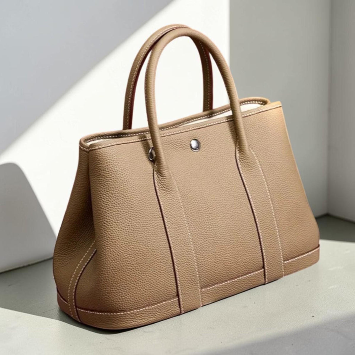 May include: A tan leather tote bag with two top handles and a silver-tone closure. The bag features white stitching and a structured design, suitable for everyday use.