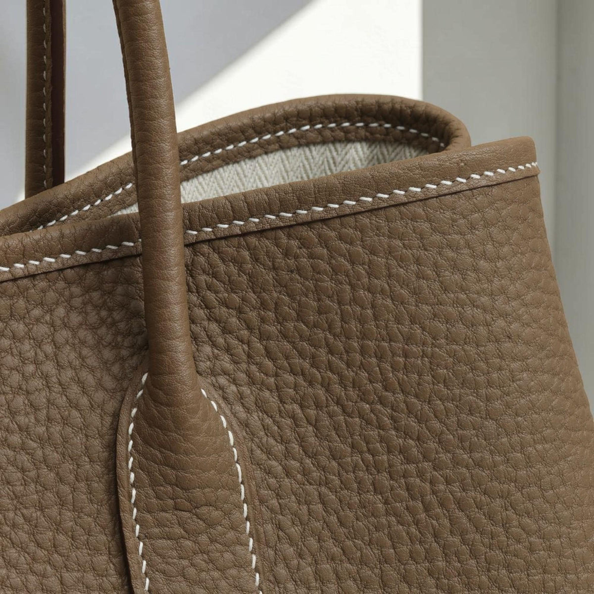 May include: Close-up of a brown leather tote bag with a textured surface. The bag features a sturdy handle and white stitching along the edges. The bag's interior is visible, showcasing a light-colored fabric.