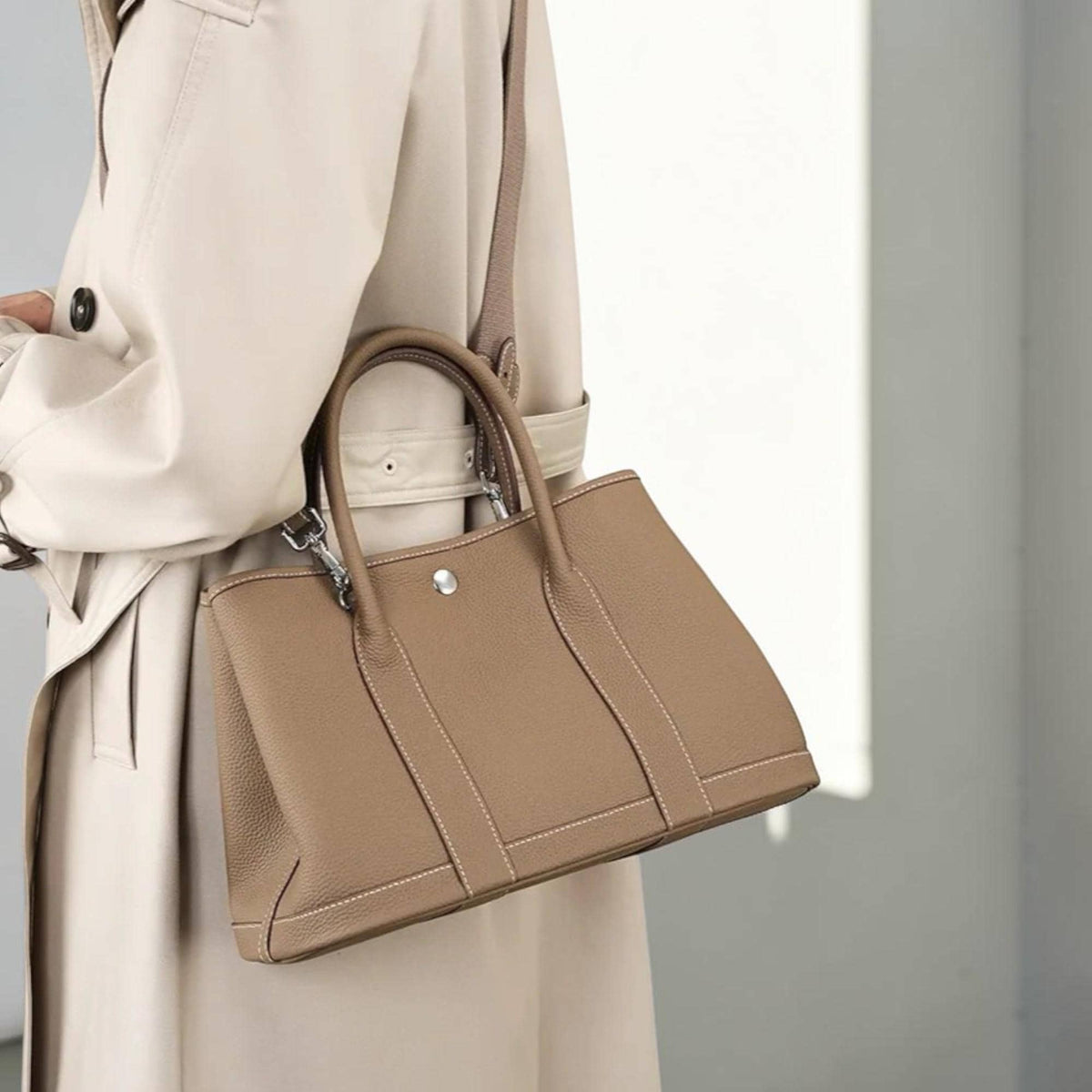 May include: A tan leather tote bag with a structured design. The bag features a top handle and a detachable shoulder strap. White stitching accents the edges, and a silver-tone clasp secures the top. The bag is shown against a neutral background.