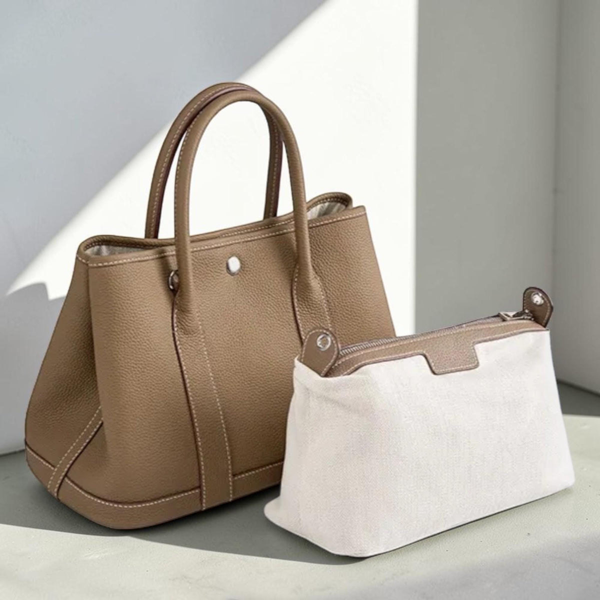 May include: A taupe leather tote bag with a matching small pouch. The tote has two handles, a silver button closure, and white stitching. The pouch is white canvas with a brown leather trim and a zipper closure.