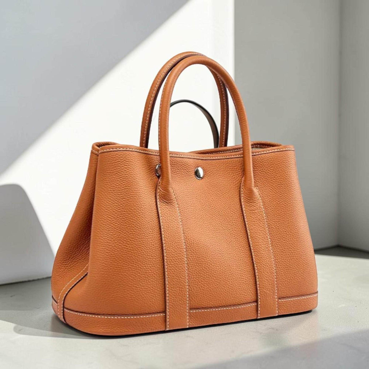May include: A tan leather tote bag with two top handles and a silver-tone clasp. The bag features white stitching along the seams and a structured design. The bag is sitting on a light-colored surface.